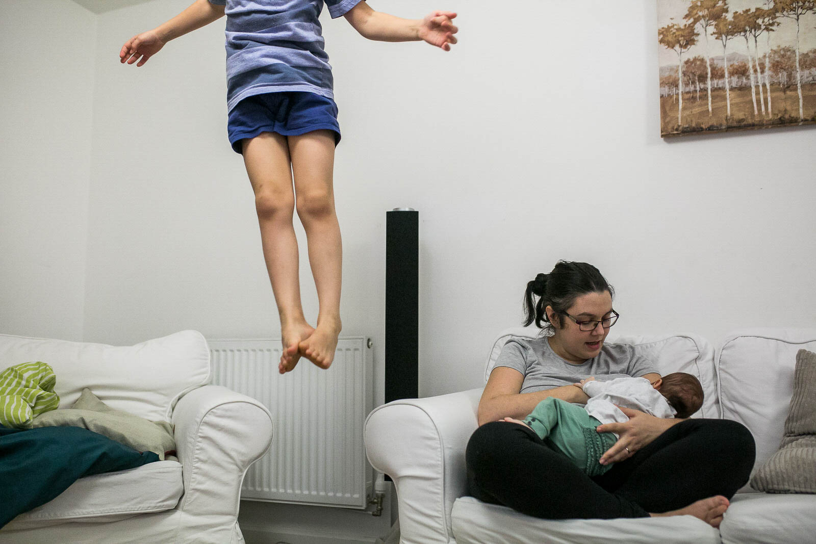 Young child leaping off the sofa while mother is breastfeeding a baby during a family photoshoot in London.